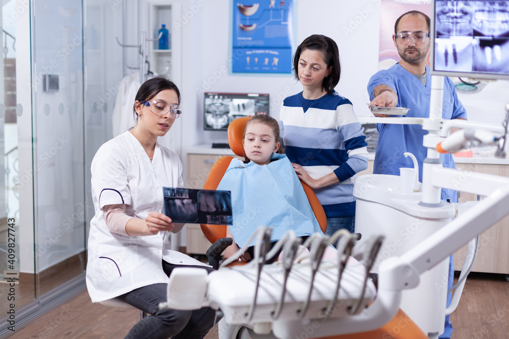 Dentist talking about tooth procedure holds radiography talking with little kid. Stomatologist explaining teeth diagnosis to mother of child in health clinic holding x-ray. Dentist talking about tooth procedure holds radiography talking with little kid. Stomatologist explaining teeth diagnosis to mother of child in health clinic holding x-ray.