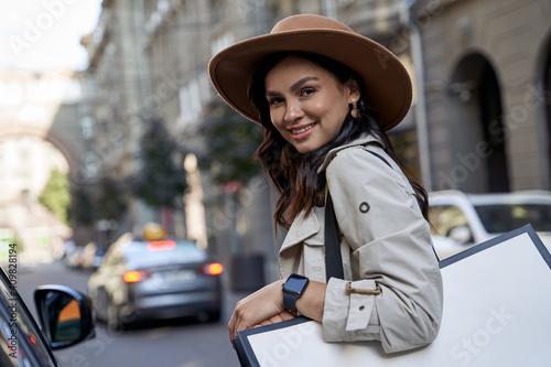 Feeling happy after shopping. Portrait of a young beautiful caucasian woman in hat with shopping bag looking at camera and smiling while standing on the city street