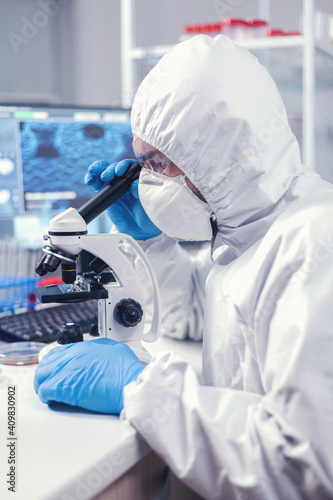 Researcher in modern microbiology laboratory uses microscope dressed in ppe suit. Virolog in coverall during coronavirus outbreak conducting healthcare scientific analysis. photo