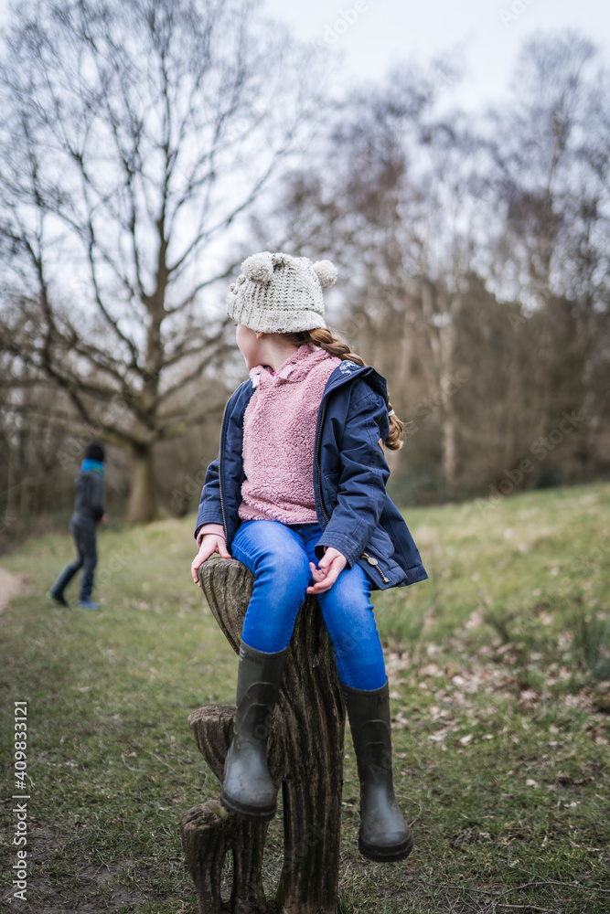 Fototapeta premium Excited happy young girl in wellinton boots sat in autumn countryside looking back away from camera at boy exploring the great outdoors nature park with bobble hat pink jumper and blue coat in winter