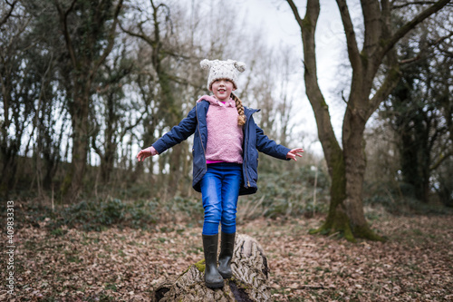 Excited happy young girl in wellinton boots sat in autumn countryside balancing on tree log with arms out exploring the great outdoors nature park with bobble hat pink jumper and blue coat in winter