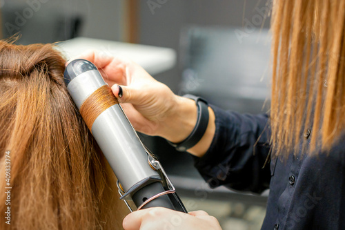Professional hairdresser makes curls with a curling iron for a young woman with long red hair in a beauty salon