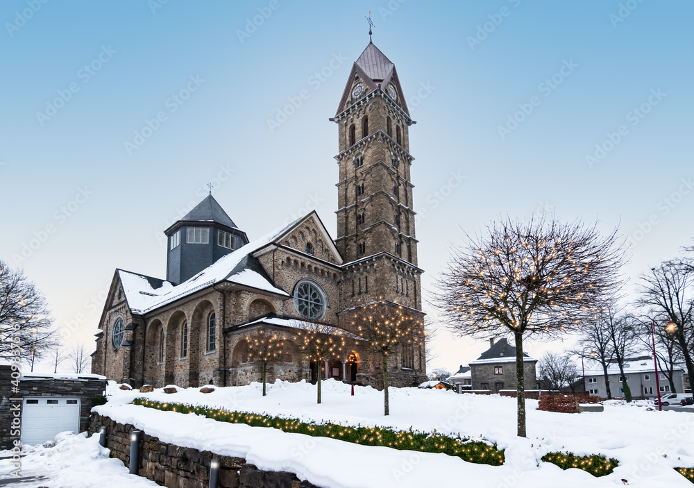 Beautiful church in Butgenbach surrounded by a winter landscape with illuminated Christmas lights in the trees around Christmas time. 