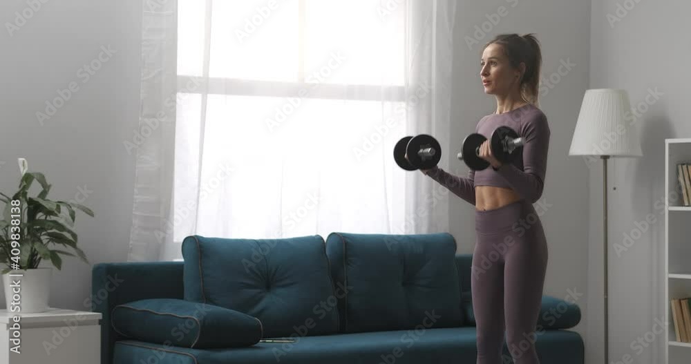 pretty sportswoman is training with dumbbells in apartment at daytime, full-length portrait of athletic lady in apartment with modern interior