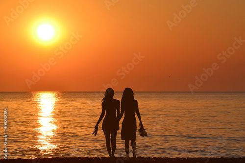 Two  young women holding hands walking barefoot with slippers in his hand on the beach