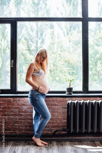 Young pregnant woman standing near window at home. Happy expectant mother thinking about her baby, grooming her belly. The last months of pregnancy. The concept of pregnancy, rest and expectation.