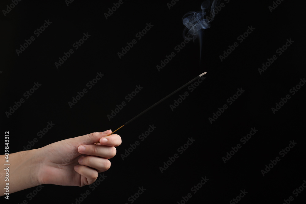 Woman holding smoldering incense stick on black background, closeup. Space for text