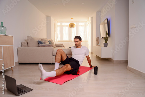 Young man massages hamstrings by foam roller at home in front of a laptop. Online workout. Home training concept.