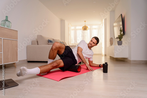 Young sporty man doing Side Planking with Foam Rollerr at home in front of a laptop. Online workout. Home training concept.