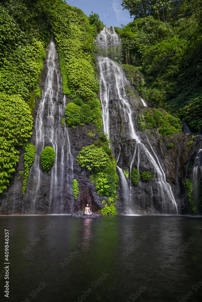 Fototapeta premium Yoga lotus pose. Young Caucasian woman sitting on the stone, meditating, practicing yoga, pranayama at waterfall. Hands in gyan mudra. Yoga retreat. Copy space. Banyumala waterfall, Bali