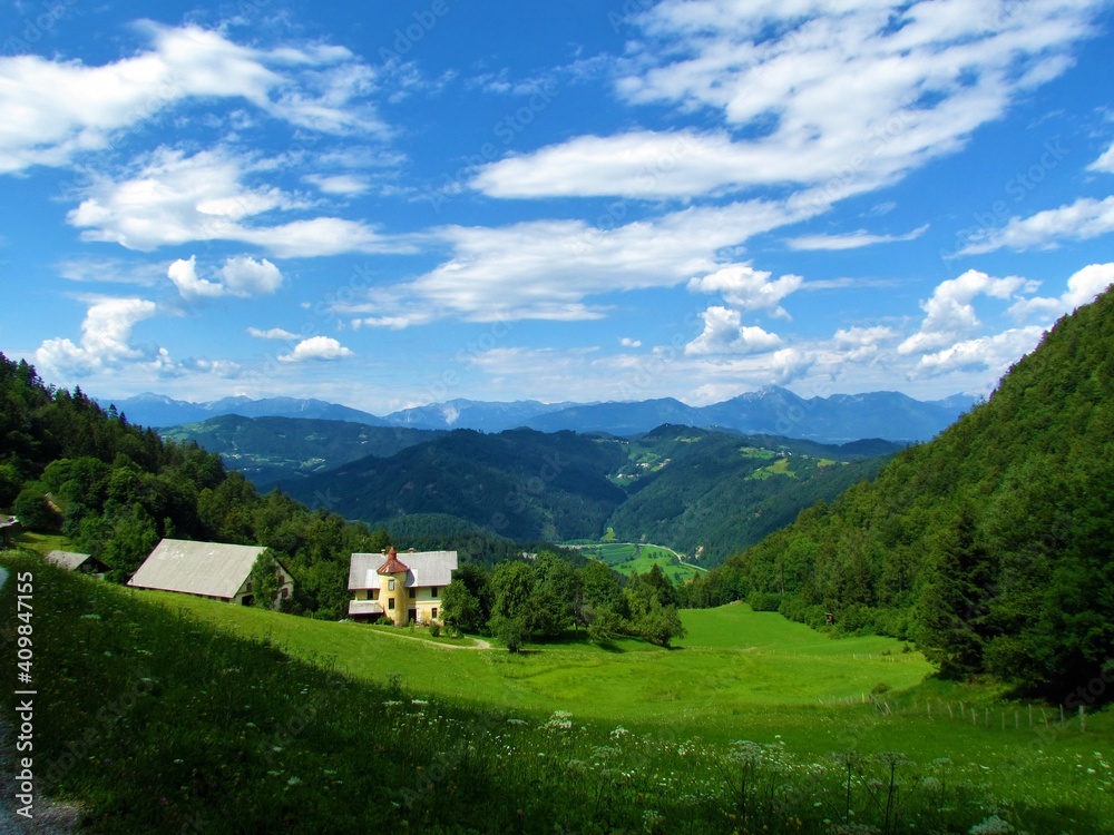 view of hills and mountains of Gorenjska region of Slovenia with a large meadow surrounded by forest and a old rustic farmhouse with a tower