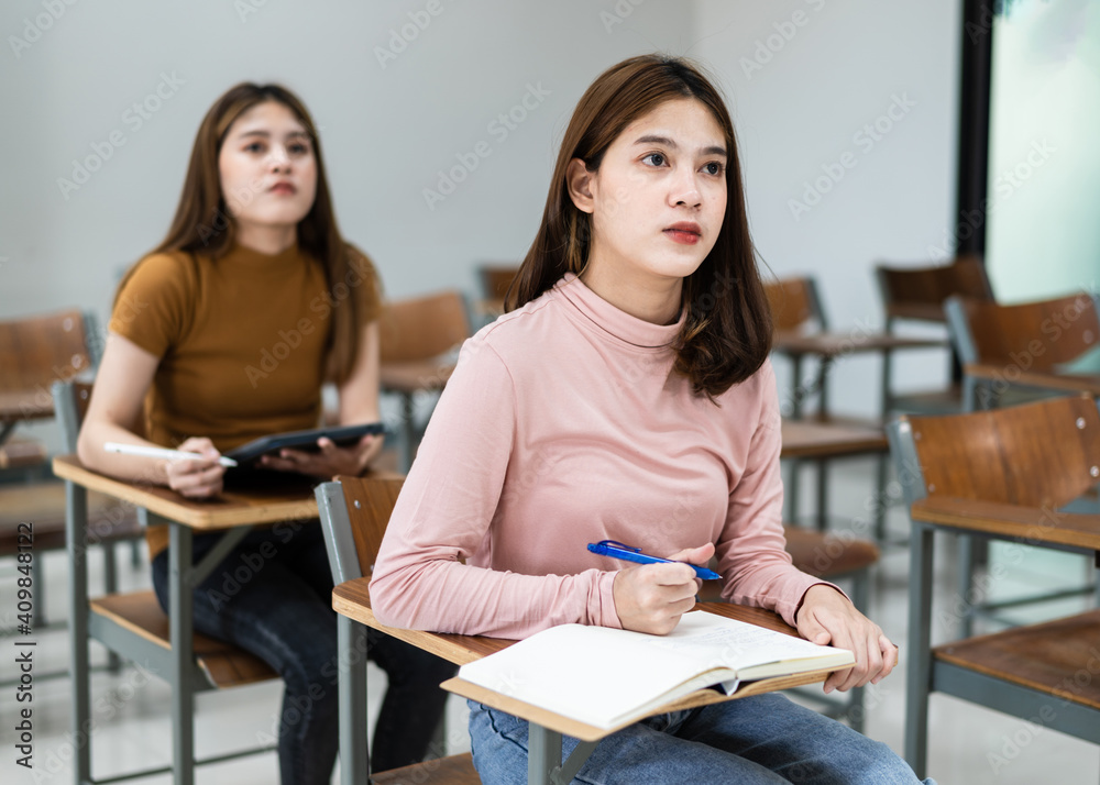 Selective focus of the teen college students sitting on lecture chair in classroom write on examination paper answer sheet in doing the final examination test. Female students in the student uniform. Selective focus of the teen college students sitting on lecture chair in classroom write on examination paper answer sheet in doing the final examination test. Female students in the student uniform.