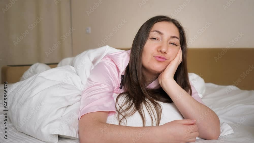 Nice brunette woman in pajama smiles gently and sincerely showing air kisses to coming husband lying on king size bed in bedroom close view Nice brunette woman in pajama smiles gently and sincerely showing air kisses to coming husband lying on king size bed in bedroom close view