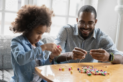 Close up smiling African American father and little daughter playing with colorful beads, sitting on couch at home, crafting bracelet, family involved in creative activity, enjoying leisure time