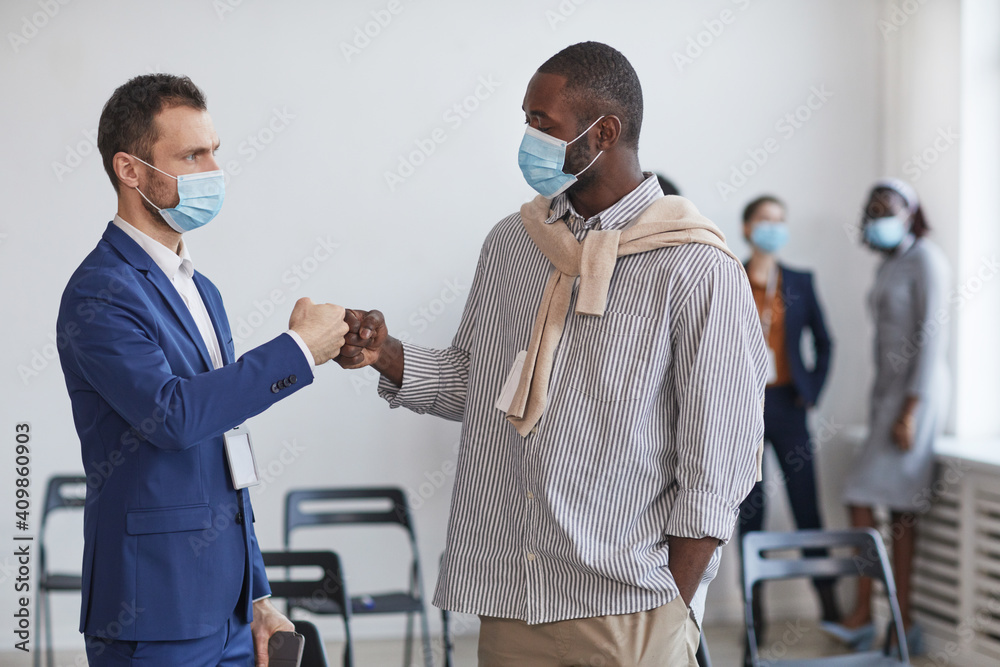Obraz premium Waist up portrait of two businessmen wearing masks and bumping fists as contactless greeting during break at business conference, copy space