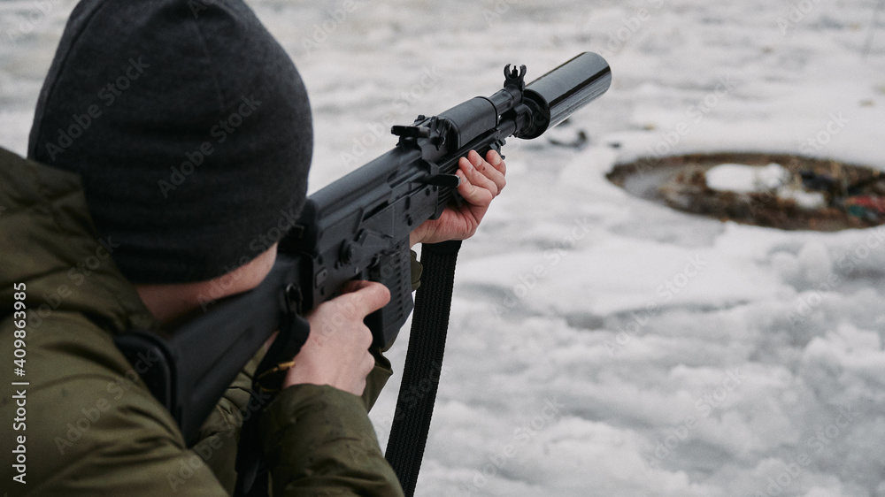 A shooter in a cap and green jacket aiming at a target with a civilian Kalashnikov-type machine gun, with a silencer in winter A shooter in a cap and green jacket aiming at a target with a civilian Kalashnikov-type machine gun, with a silencer in winter