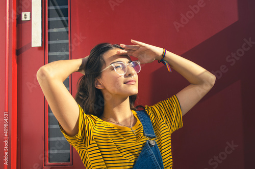 Portrait of a beautiful caucasian teenager girl wearing glasses and colorful clothes with her right hand behind her head and left hand over her eyes. Dark red door on the background. Copy space.