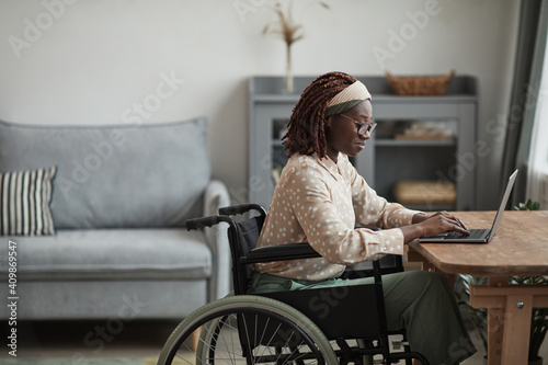 Side view portrait of young African-American woman using wheelchair while working from home in minimal grey interior, copy space photo