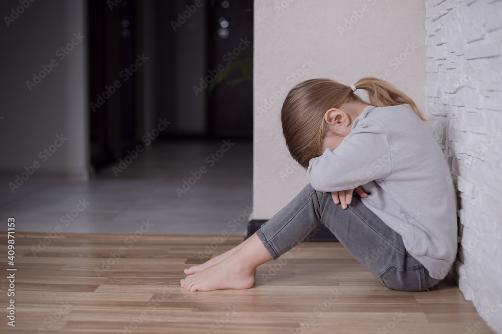 Sad little child girl sitting on floor in corner at home. Stock Photo