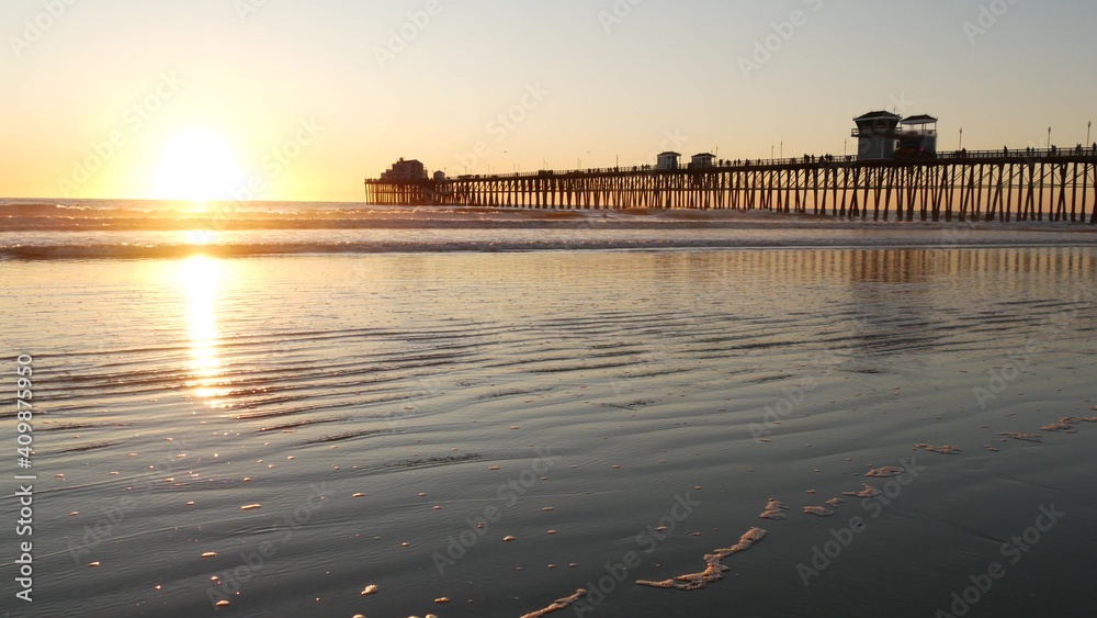 Fototapeta premium Wooden pier on piles, silhouette at sunset, California USA, Oceanside. Waterfront resort, pacific ocean tide, tropical beach. Summertime coastline vacations atmosphere. Sunny sea waves at sundown.