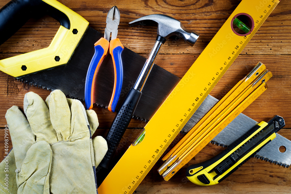 Handsaw, hammer, protective gloves, level, model knife, folding ruler and pliers on wooden background Handsaw, hammer, protective gloves, level, model knife, folding ruler and pliers on wooden background