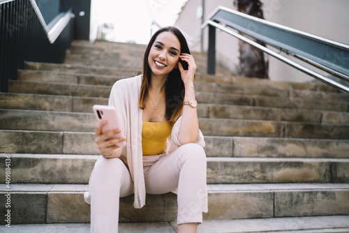 Portrait of cheerful hipster girl holding modern mobile technology for phoning during leisure time in city, toothy Caucaisan female blogger with smartphone gadget in hand smiling at camera while rest