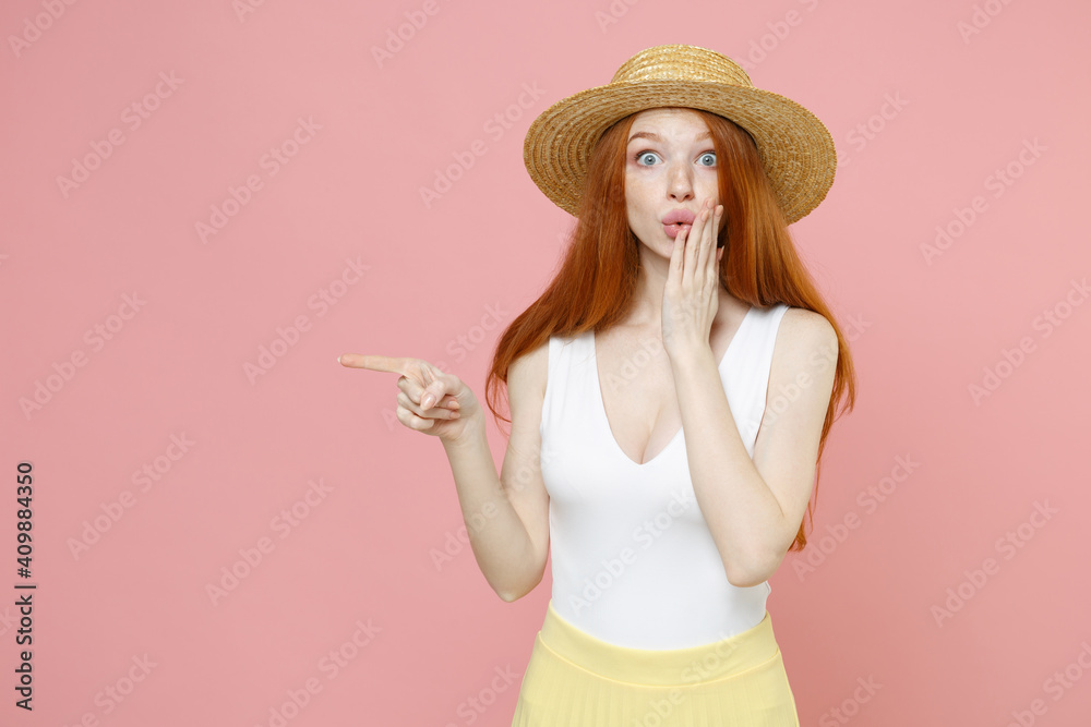 Young surprised shocked redhead woman 20s ginger long hair wear straw hat summer clothes pointing finger aside on copy space workspace area mock up isolated on pastel pink background studio portrait