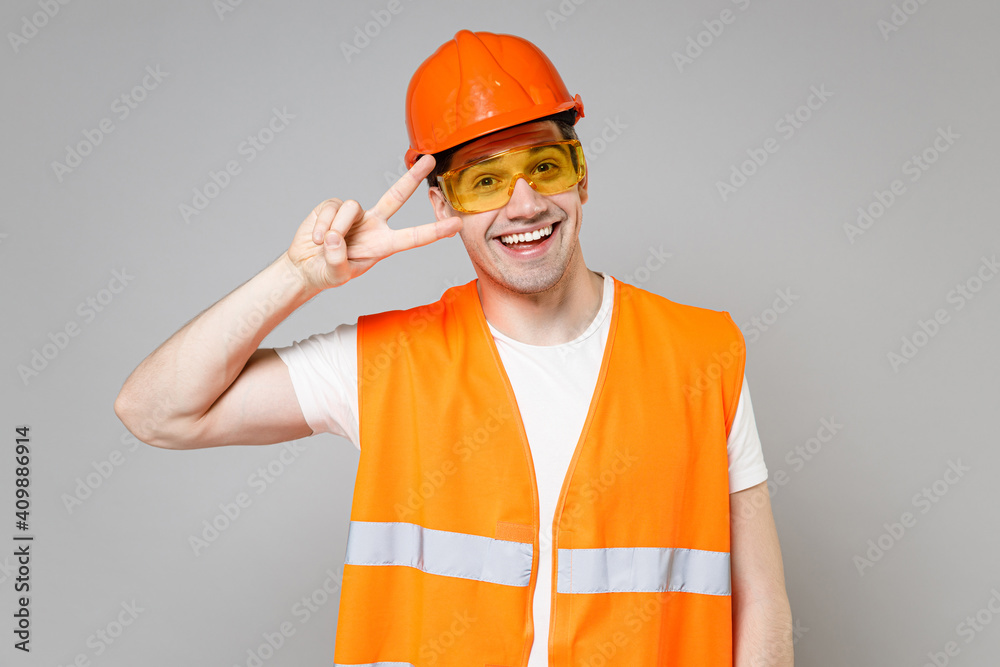 Young employee friendly handyman man in orange vest protective hardhat victory v-sign gesture isolated on grey background studio portrait Instruments for renovation apartment room Repair home concept