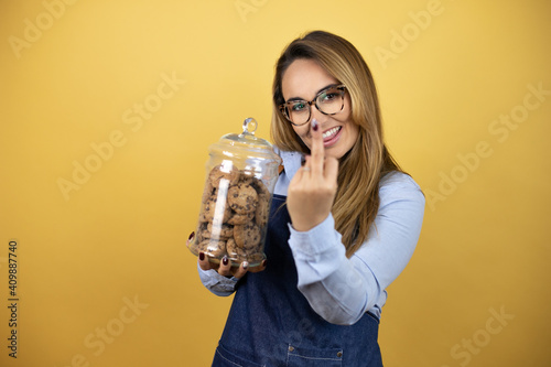 Young hispanic woman wearing baker uniform holding a cookies jar showing middle finger doing fuck you bad expression, provocation and rude attitude. screaming excited