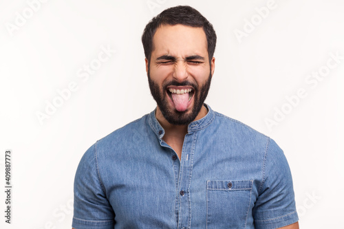 Naughty infantile man with beard closing eyes and showing tongue, having fun, fooling around, childish manners. Indoor studio shot isolated on white background © khosrork