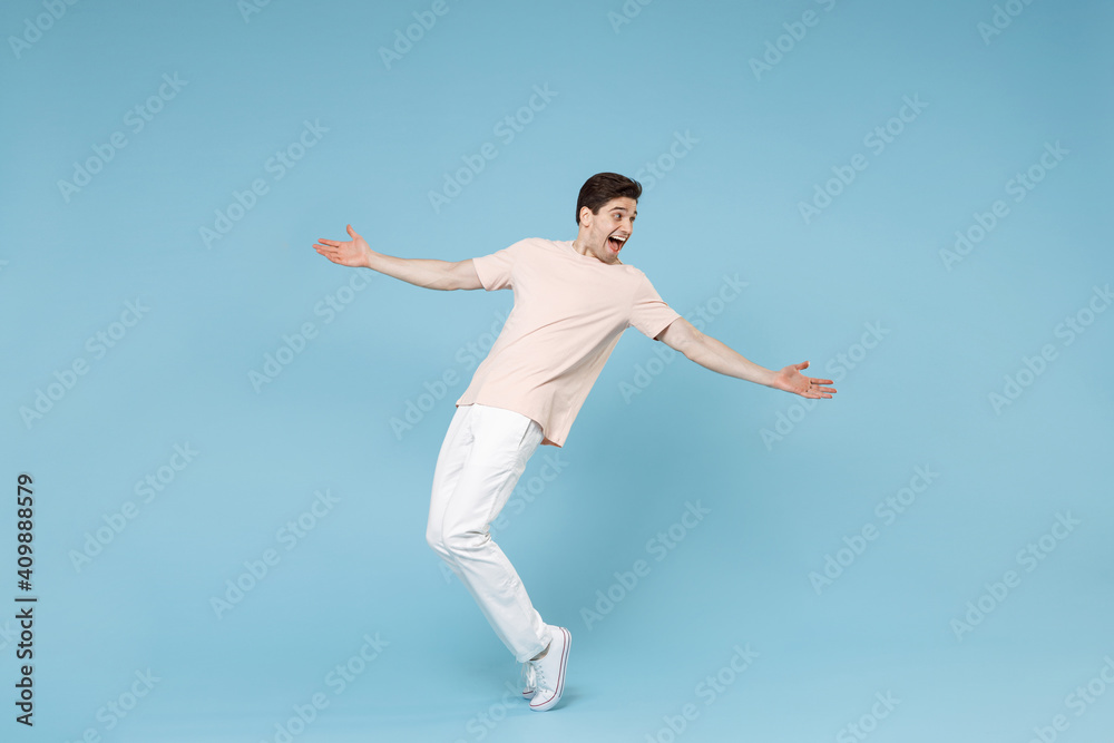 Full length of young caucasian attractive smiling student man 20s in beige t-shirt white pants leaning over with outstretched hands looking aside isolated on blue color background studio portrait