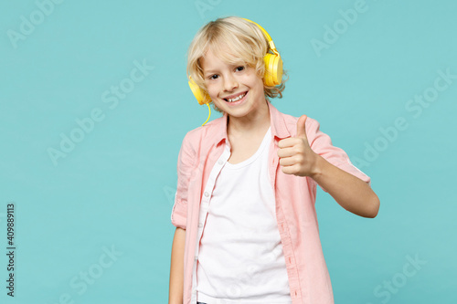 Smiling little curly kid boy 10s years old in pink shirt listening music with headphones showing thumb up isolated on blue turquoise background children studio portrait. Childhood lifestyle concept.