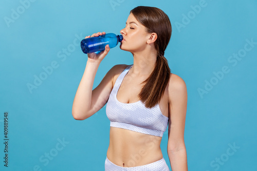 Side view thirsty athletic woman in white sportswear drinking spring water from bottle, restoring water balance after exhausted workout. Indoor studio shot isolated on blue background photo