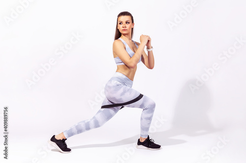 Side view full length assertive concentrated woman gymnast in white top and tights doing fitness exercises using elastic band, pumping legs and buttocks. Indoor studio shot isolated on gray background