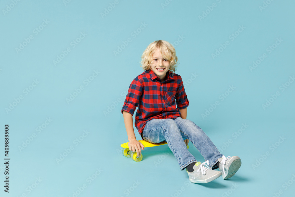 Full length of smiling little curly kid boy 10s in basic red checkered shirt sitting on skateboard looking camera isolated on blue background children studio portrait. Childhood lifestyle concept. Full length of smiling little curly kid boy 10s in basic red checkered shirt sitting on skateboard looking camera isolated on blue background children studio portrait. Childhood lifestyle concept.