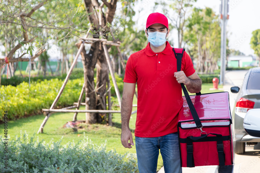Portrait Smiling young delivery man in red uniform holding a box in front of the house. Protection face mask quarantine pandemic coronavirus virus 2019-ncov concept
