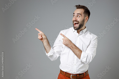 Surprised young bearded business man in classic white shirt standing point index fingers aside up on mock up copy space isolated on grey background studio portrait. Achievement career wealth concept.