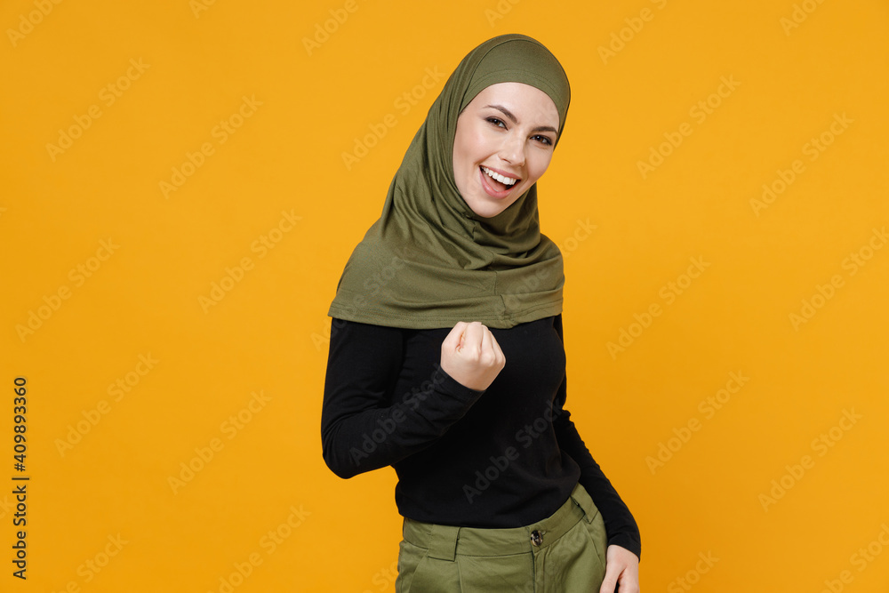 Funny young arabian muslim woman in hijab black green clothes doing winner gesture celebrating clenching fist say yes isolated on yellow background studio portrait. People religious lifestyle concept. Funny young arabian muslim woman in hijab black green clothes doing winner gesture celebrating clenching fist say yes isolated on yellow background studio portrait. People religious lifestyle concept.