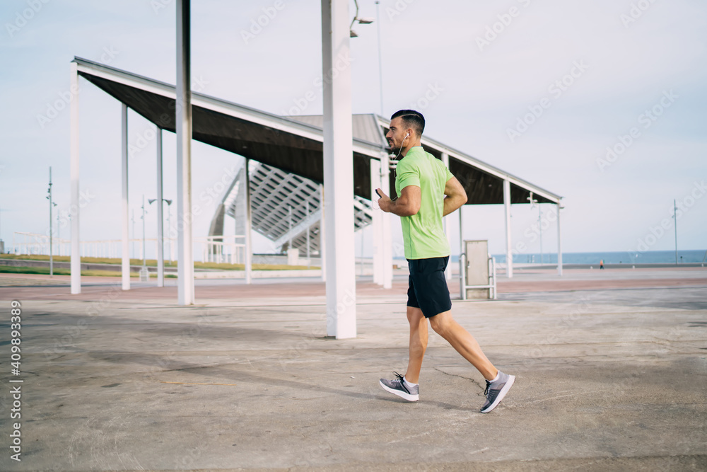 Side view of determined male athlete in electronic headphones listening music songs during cardio running around city, Caucaisan sportsman in modern earphones running during physical recreation