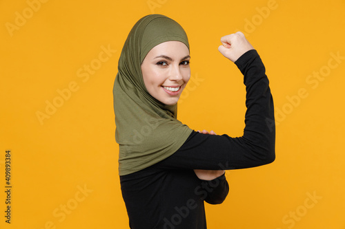 Fototapeta Side view of funny sporty young arabian muslim woman in hijab black green clothes showing biceps muscles on hands isolated on yellow background, studio portrait