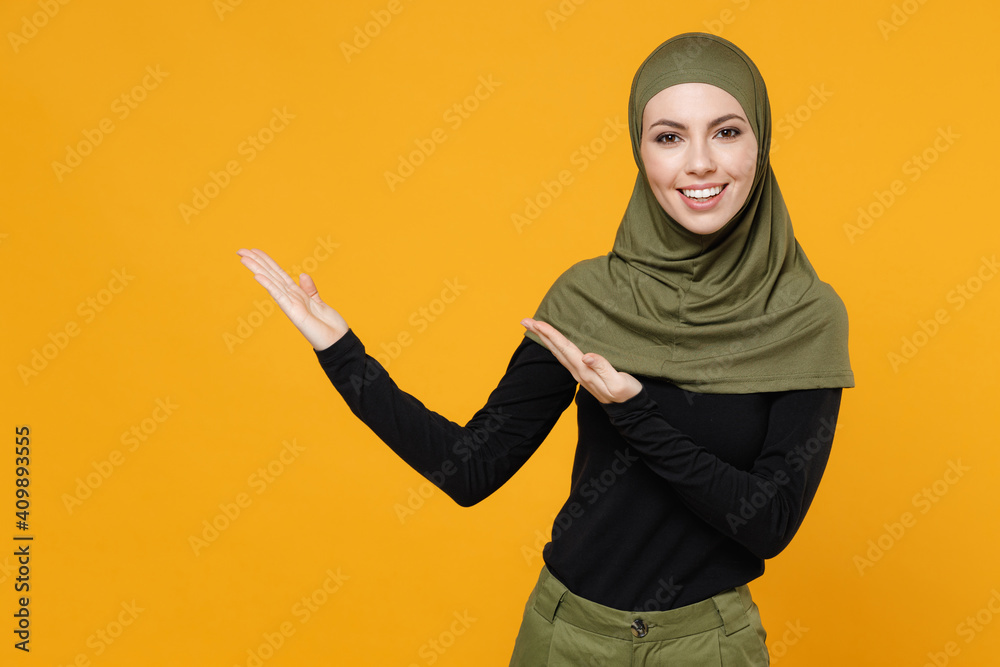Cheerful young arabian muslim woman in hijab black green clothes pointing hands aside on mock up copy space isolated on yellow color background, studio portrait. People religious lifestyle concept. Cheerful young arabian muslim woman in hijab black green clothes pointing hands aside on mock up copy space isolated on yellow color background, studio portrait. People religious lifestyle concept.