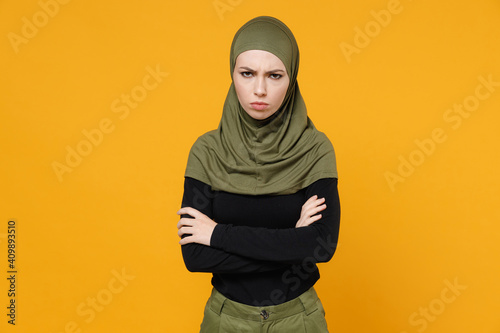 Displeased upset young arabian muslim woman in hijab black green clothes holding hands crossed looking camera isolated on yellow color background, studio portrait. People religious lifestyle concept.