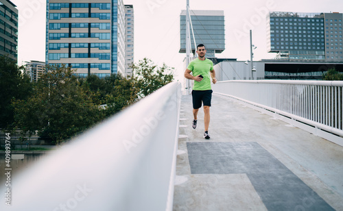 Caucasian sportsman in electronic earphones using smartphone media application for listening music playlist and running around city streets, muscular male bodybuilder with cellphone jogging © BullRun