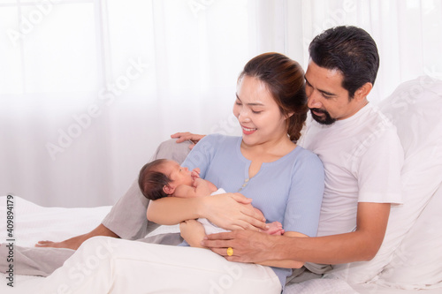 Beautiful wife leaning her back to husband leg while hold newborn with love and carefully, happy Hispanic father with beard or mix race hug mother and first child son on white bed, family concept