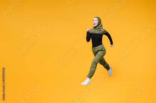 Full length side view of excited young arabian muslim woman in hijab black green clothes jumping like running isolated on yellow color background studio portrait. People religious lifestyle concept.