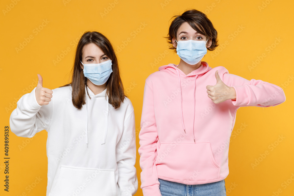 Cheerful funny two young women friends 20s in casual hoodies sterile face mask to safe from coronavirus virus covid-19 showing thumbs up isolated on bright yellow wall background studio portrait.