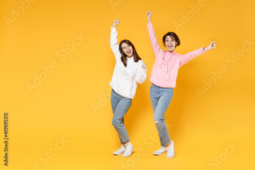 Full length of joyful excited two young women friends 20s wearing basic white pink hoodies doing winner gesture celebrating clenching fists say yes isolated on yellow color background studio portrait.