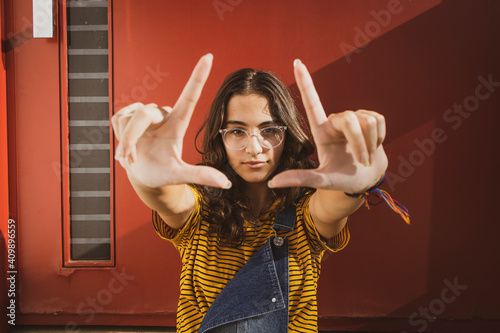 Portrait of a teenager girl wearing clear transparent glasses and colorful clothes making frame formation with hands against red door. photo