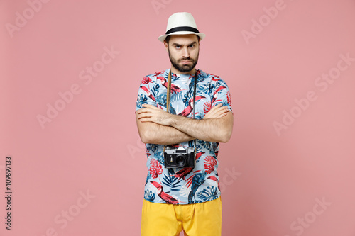 Dissatisfied young traveler tourist man in summer casual clothes hat with photo camera holding hands crossed isolated on pink background. Passenger traveling on weekends. Air flight journey concept.