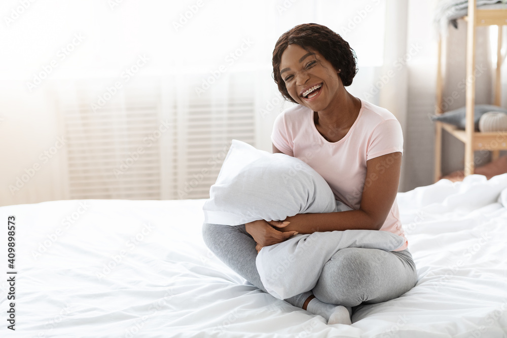 Laughing black woman in pajamas sitting on bed, hugging pillow